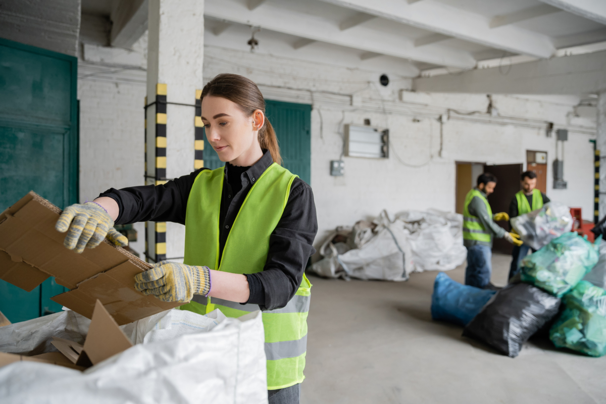 Young Female Worker In Protective Vest And Gloves Holding Cardboard Near Sack And Blurred Colleagues Working In Waste Disposal Station, Garbage Sorting And Recycling Concept